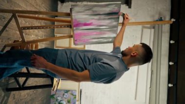 Bir sanat atölyesinde dikey video görüntüsü. Ressam tuvale pembe damlalar serpiştiriyor.