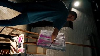 Karanlık bir sanat stüdyosunda dikey bir video mavi tişörtlü bir sanatçı tuval üzerinde fırçayla resim yapıyor.
