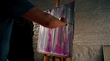 Karanlık bir sanat stüdyosunda mavi tişörtlü bir sanatçı tuvalde fırçayla resim yapıyor.
