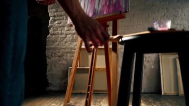 Karanlık bir sanat stüdyosunda bir sanatçı palete palet bıçağı siler ve başka bir palet alır.