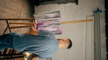 Genel olarak bir sanat stüdyosunda çekilen dikey video, sanatçı tuvalde geniş bir fırçayla çizgiler oluşturur.