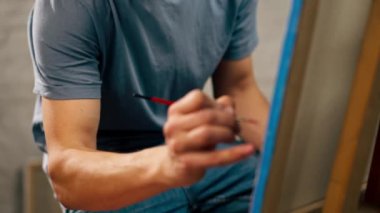 Bir sanat atölyesinde mavi tişörtlü genç bir sanatçı otururken bir resim üzerinde çalışıyor.