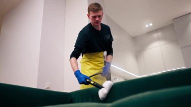 Sarı önlüklü bir apartman temizleyicisinde profesyonel ekipmanla yeşil bir kanepeyi buharla temizliyor.