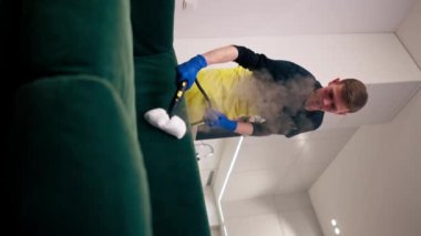 Sarı önlüklü bir apartman temizleyicisindeki dikey video profesyonel ekipmanla yeşil bir kanepenin buharlı temizliğini yapıyor.