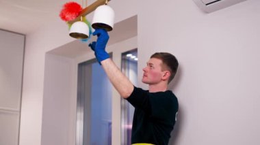 Apartmanda sarı önlüklü bir temizlikçi avizeyi toz fırçasıyla temizliyor.