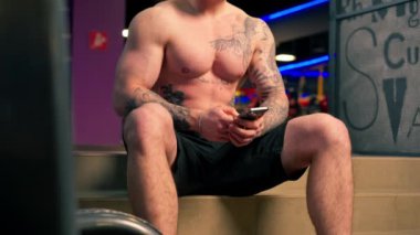 Spor salonuna yakın dövmeli genç yakışıklı bir adam oturuyor ve telefonda bir mesaj okuyor.