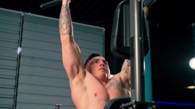 Spor salonunda dövmeli genç bir adam barfiks çekiyor. Sırtını ve kollarını sallıyor.