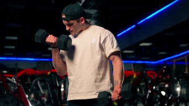 Spor salonunda tişörtlü ve şapkalı genç yakışıklı bir adam dambıllarla egzersiz yapıyor ve kollarını sallıyor.