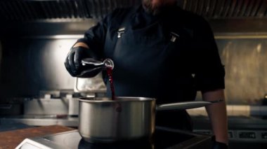 Profesyonel mutfakta siyah ceketli bir şef ocağın üzerinde duran tencereye kırmızı sos döker.