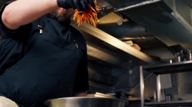 Profesyonel bir mutfakta, siyah eldivenler giyip, demir bir kaseye havuç dolduruyor.