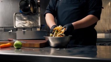 Profesyonel bir mutfakta, siyah eldivenlerle, doğranmış sebzeleri demir bir kasede karıştırarak.