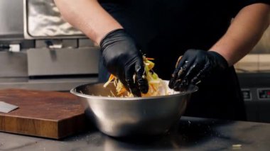 Profesyonel bir mutfakta, siyah eldivenlerle, doğranmış sebzeleri demir bir kasede karıştırarak.