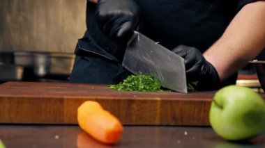 Profesyonel bir mutfakta siyah ceketli bir şef masada maydanoz doğruyor.