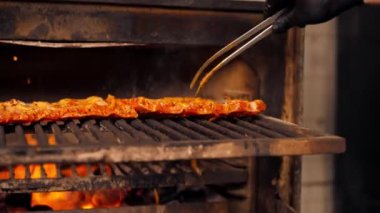 Profesyonel bir mutfakta siyah ceketli bir şef kızgın ızgara fırınının yanında marine edilmiş pirzola serer.