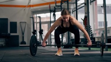 Spor salonunda siyah elbiseli bir koç kız halter ve ağırlık egzersizleri yapıyor.