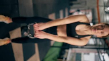 Spor salonundaki dikey video siyah taytlı bir kız antrenör ve bir top ağırlık ile çalışıyor.