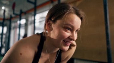 Spor salonunda bir kız antrenör bir eliyle dambılı kaldırıyor ve gülümsüyor.