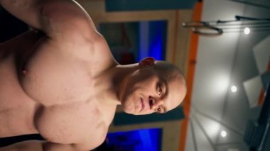 Spor salonunda genç, yakışıklı ve kel bir antrenör iki ağır ağırlıkla çalışıyor ve kollarını kaldırıyor.