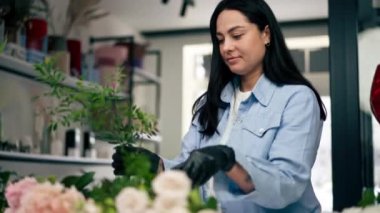 Bir çiçekçide, beyaz bir masanın yanındaki bir kız bir sepet çiçeği süslemek için zeytin ağacının dalını buduyor.