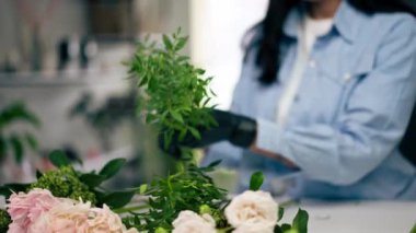 Bir çiçekçide, beyaz bir masanın yanında bir kız bir sepet çiçeği süslemek için zeytin ağacının dalını buduyor.