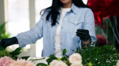 Bir çiçekçide, beyaz bir masanın yanındaki bir kız bir sepet çiçeği süslemek için zeytin ağacının dalını buduyor.