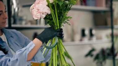 Bir çiçekçide, beyaz bir masanın üzerindeki bir kız neredeyse bitmiş bir buketi yapışkan bantla sarar.
