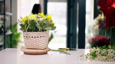Bir çiçekçide, beyaz bir masanın yanında bir kız çiçek aranjmanı topluyor sepette bir buket çiçek.