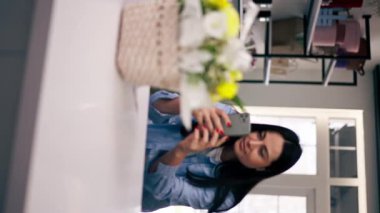 Çiçekçi dükkanındaki dikey videoda çiçekçi telefonundaki sepetin içinde toplanmış sarı bir buketin fotoğrafını çekiyor.