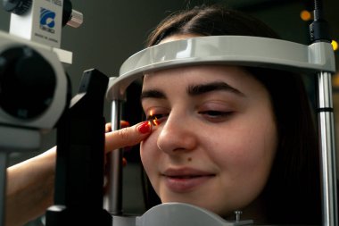 Bir göz hekimliği kliniğinde genç bir doktor büyüteçle genç bir kıza teşhis koyuyor.