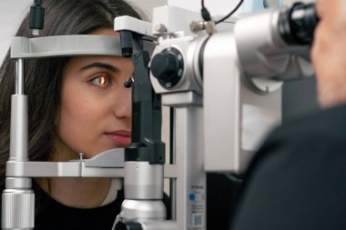 Göz hekimliği kliniğinde yaşlı bir doktor büyüteçle genç bir kıza teşhis koyuyor.