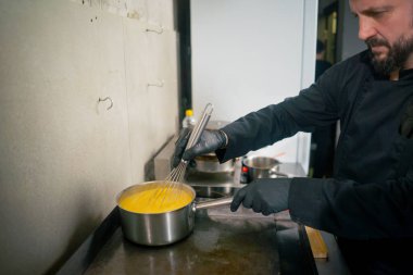 Profesyonel bir mutfakta siyah eldivenli bir şef makarna için peynir sosu pişirir.