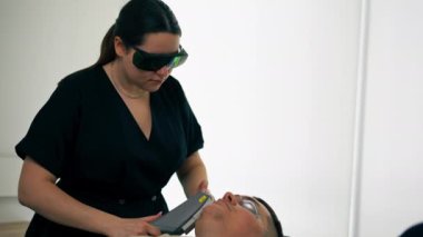 Bir kozmetoloji kliniğinde bir kız kozmetik uzmanı genç bir adamın yüzü için fotoğraf terapisi yapıyor.