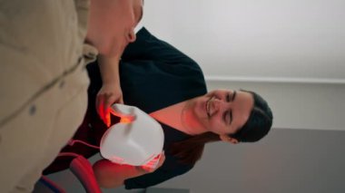 Bir kozmetoloji kliniğinde dikey video çekilirken bir kız kozmetik uzmanı bir adama nasıl fotoğraf maskesi kullanacağını söyler.