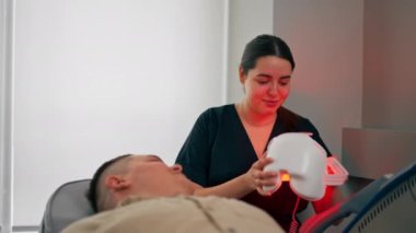 Bir kozmetoloji kliniğinde bir kız kozmetik uzmanı bir adama nasıl fotoğraf maskesi takılacağını söyler.