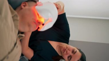 Bir kozmetoloji kliniğinde dikey video çekilirken bir kız kozmetik uzmanı bir adama nasıl fotoğraf maskesi takacağını söyler.