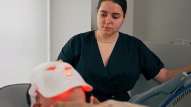 Bir kozmetoloji kliniğine yakın bir yerde bir kız kozmetik uzmanı bir adama fotoğraf maskesinin nasıl kullanılacağını söyler.