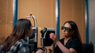 Bir göz hekiminde genç bir doktor büyüteçle genç bir kıza teşhis koyuyor.