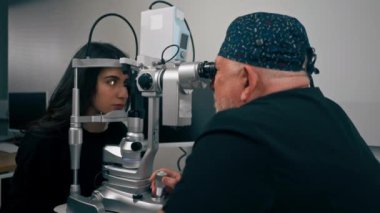 Optalmoloji kliniğinde yaşlı doktor büyüteçle genç bir kıza teşhis koyuyor.