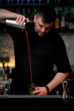 Önlüklü genç bir barmen alkol içeceği hazırlıyor. Kokteyl karıştırıcısına alkol dolduruyor.