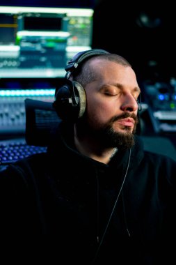 Karanlık bir kayıt stüdyosunda müzik aletlerinin ses işleme arkaplanına karşı genç bir adam oturur ve kulaklıkla müzik dinler.