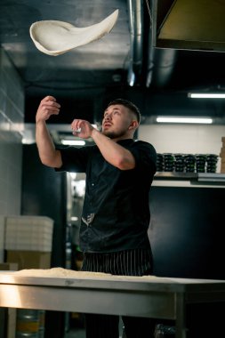 Profesyonel bir mutfakta, pizzacıda, siyah üniformalı genç bir şef havaya hamur krepi fırlatıyor.