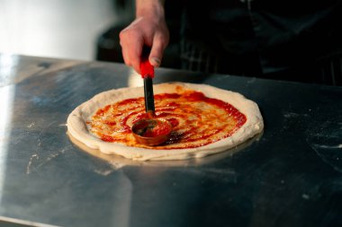 Bir pizzacıda profesyonel bir mutfakta siyah üniformalı genç bir şef pizzanın üzerine ketçap sürer.
