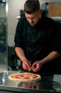 Profesyonel bir mutfakta bir pizzacıda siyah üniformalı genç bir şef pizzaya malzeme koyar.
