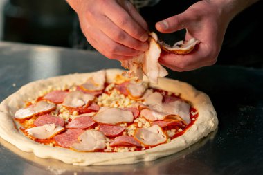 Bir pizzacının profesyonel mutfağında siyah üniformalı genç bir şef pizzanın içine malzeme koyar.