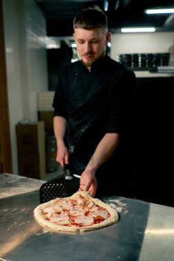 Profesyonel bir mutfakta bir pizzacıda siyahlı genç bir şef spatulayla fırın için çiğ pizza alır.