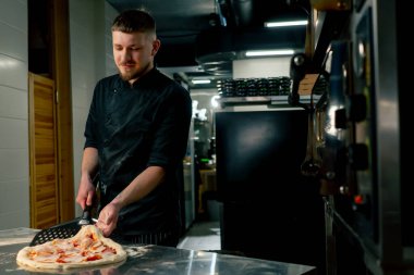 Profesyonel bir mutfakta bir pizzacıda siyahlı genç bir şef spatulayla fırın için çiğ pizza alır.
