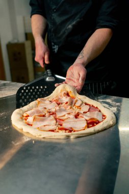 Profesyonel bir mutfakta, pizzacıda siyah giyen genç bir şef spatulayla fırın için çiğ pizza alır.