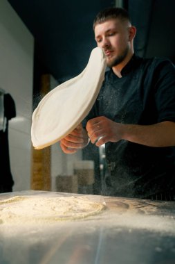 Profesyonel bir mutfakta, pizzacıda, siyah üniformalı genç bir şef havaya hamur krepi fırlatıyor.