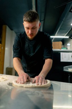 Profesyonel bir mutfakta, pizzacıda siyah üniformalı genç bir şef pizza hamuru hazırlıyor.