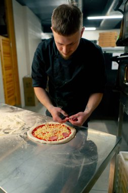 Profesyonel bir mutfakta bir pizzacıda siyah üniformalı genç bir şef pizzaya malzeme koyar.